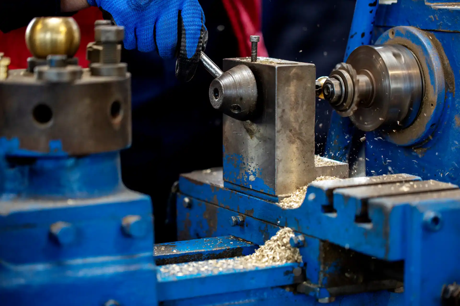 Técnico usando pistola de graxa lubrificante em uma máquina industrial de usinagem, aplicando lubrificação em uma peça metálica com luva de proteção e ambiente de oficina