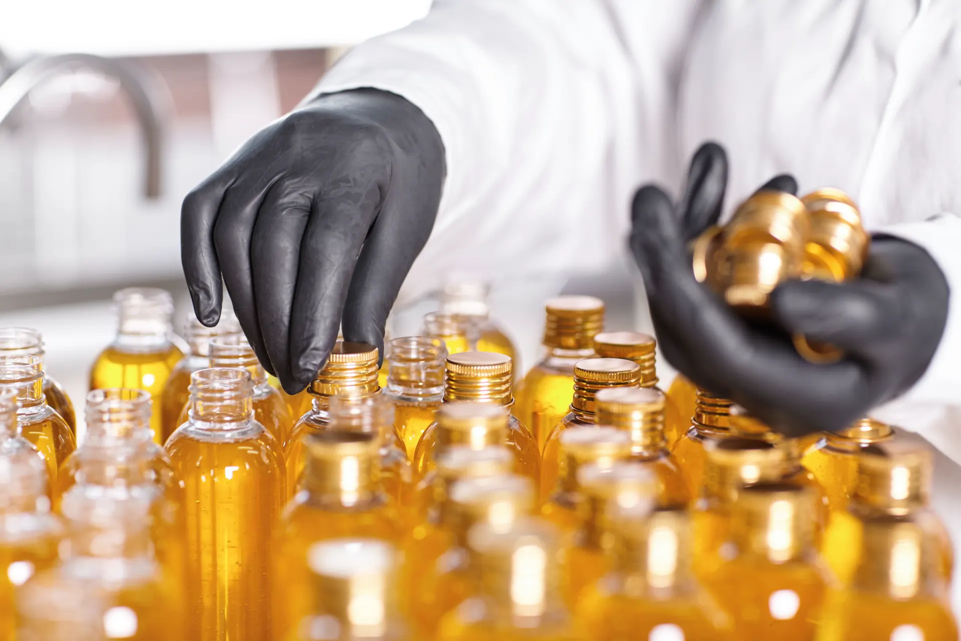 Mãos enluvadas de profissionais manipulando frascos e frascarias com líquidos âmbar, ao lado de moedas/vedações, em um cenário de controle de contaminação em ambiente de laboratório ou farmácia.